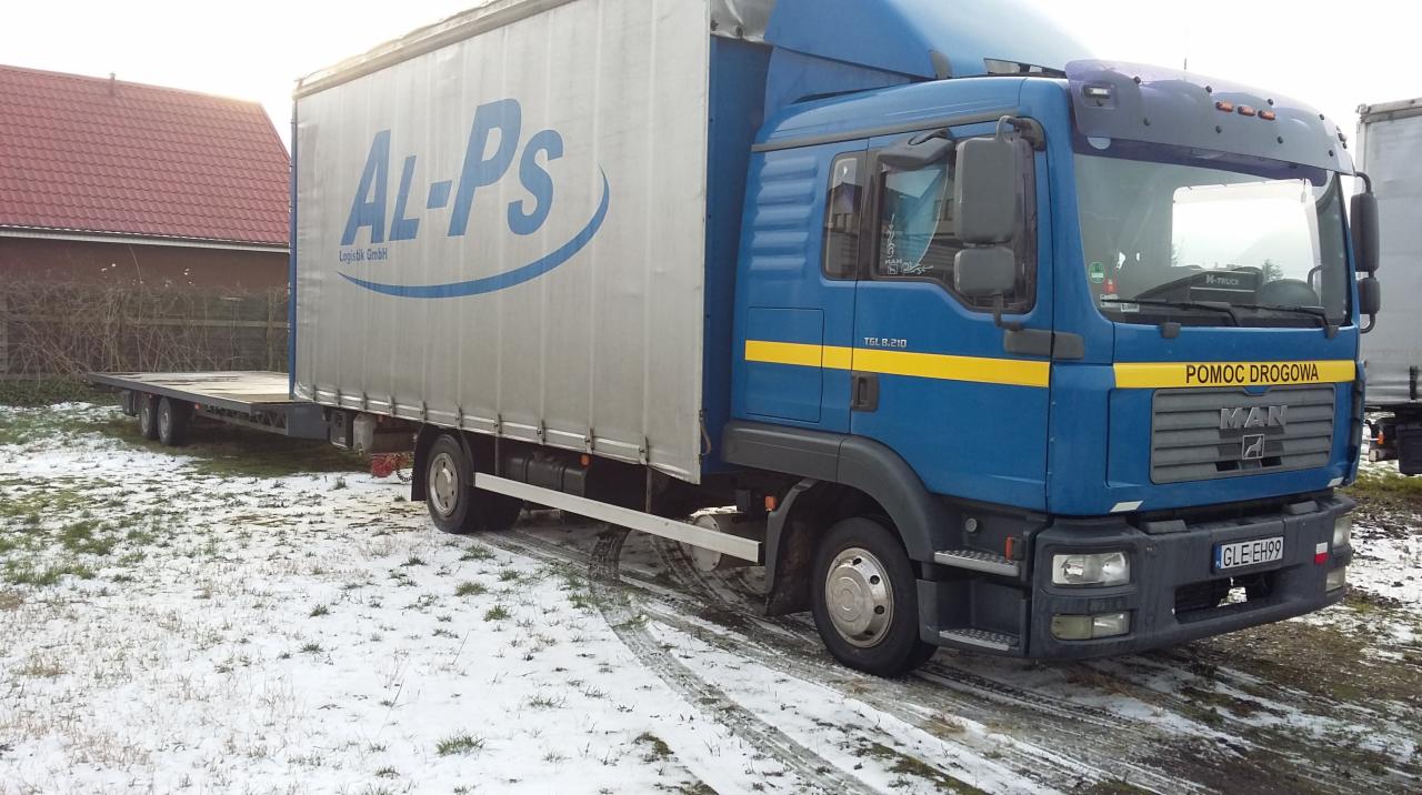 Niebieska ciężarówka MAN TGL 8.210 z naczepą platformową, oznaczona logo Al-Ps Logistik GmbH, zaparkowana na śniegu z widocznym napisem 'Pomoc Drogowa' na przedniej masce.