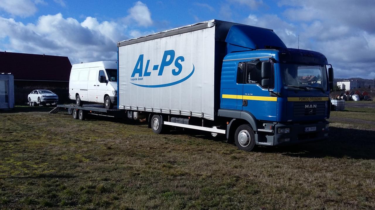 Niebieska ciężarówka MAN z naczepą marki AL-PS przewożąca białego busa na lawecie, na trawiastym placu, w tle budynki i pochmurne niebo.