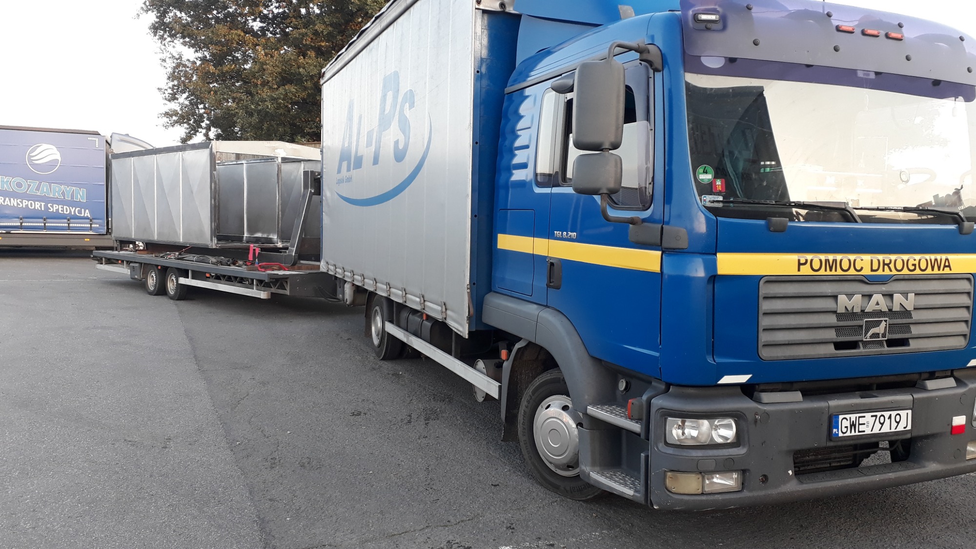 Niebieska ciężarówka MAN z napisem 'POMOC DROGOWA' i przyczepą z metalowymi kontenerami na parkingu, widoczny numer rejestracyjny GWE 7919J.