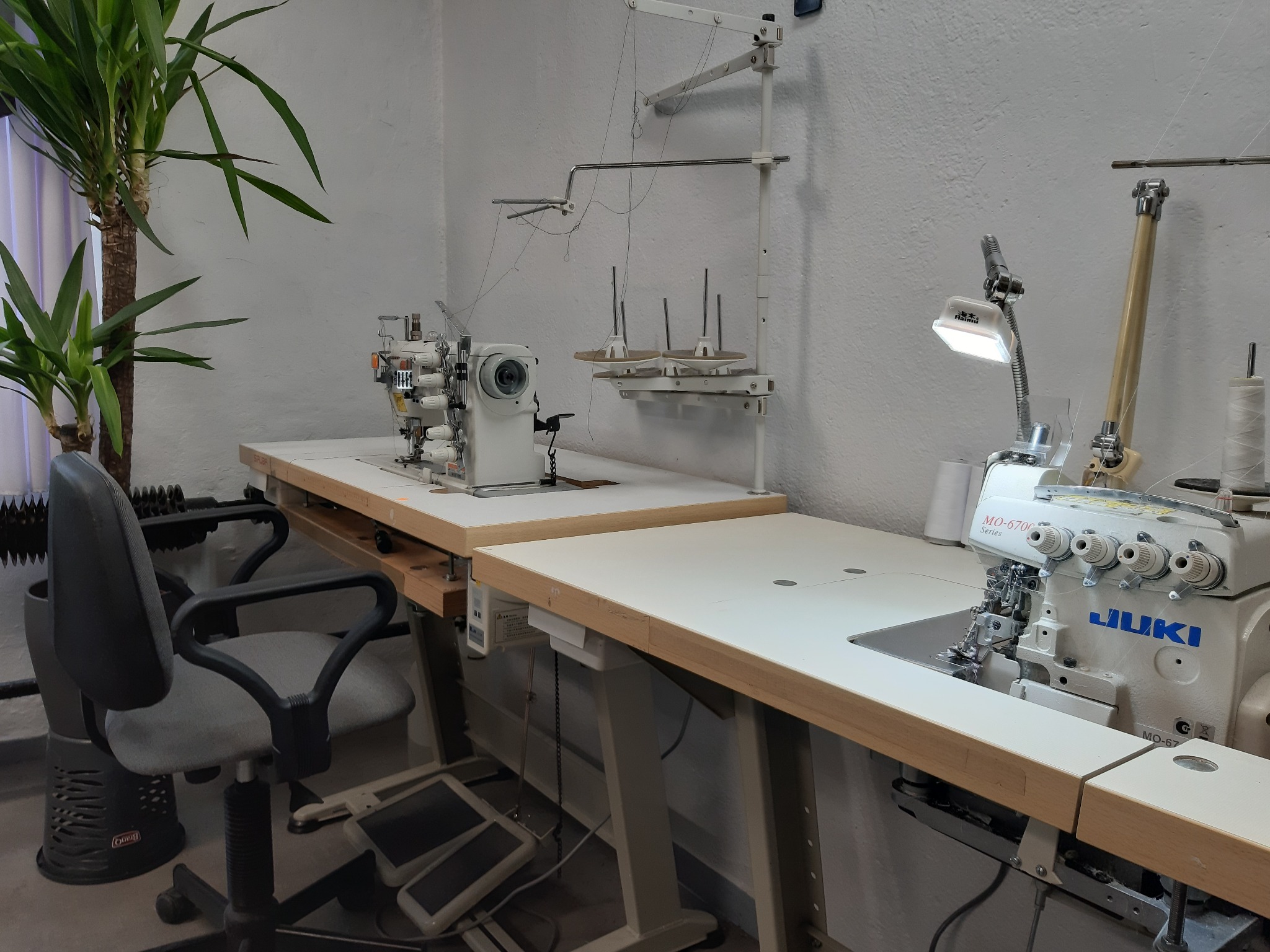 Dwie maszyny do szycia, w tym overlock JUKI MO-6700, na białych stołach w pracowni, z rośliną doniczkową w tle.