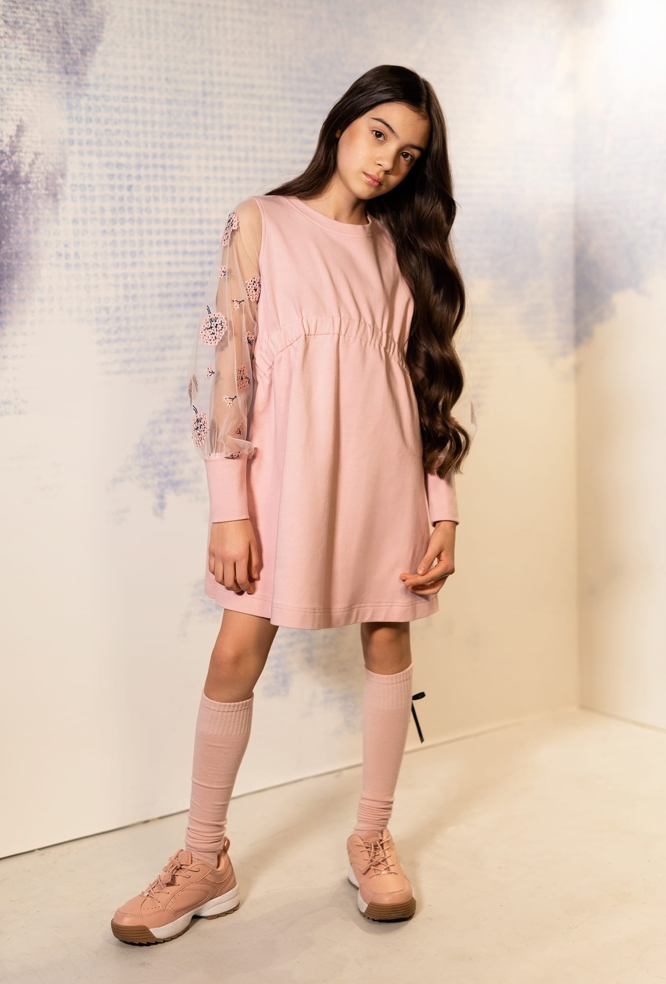 Modelka prezentuje pudrowo-różową sukienkę z transparentnymi rękawami zdobionymi aplikacjami kwiatowymi, dopełnioną podkolanówkami i sportowymi butami w tonacji.