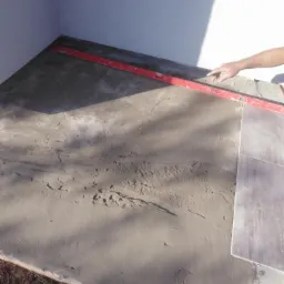 Przygotowanie podłoża pod płytki: wyrównywanie zaprawy poziomnicą, widoczny fragment ułożonej płytki i surowy teren w tle.