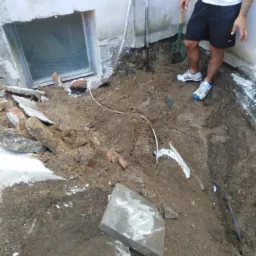 Wykop ziemi przy ścianie budynku z oknem piwnicznym, widoczne fragmenty gruzu i instalacji, mężczyzna z łopatą w tle.