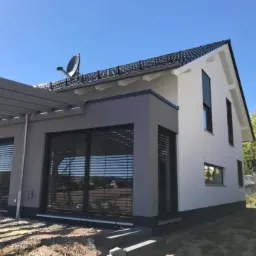 Nowoczesny dom jednorodzinny z dużymi oknami PCV z zewnętrznymi żaluzjami, widoczne rusztowanie pergoli i antena satelitarna na dachu.