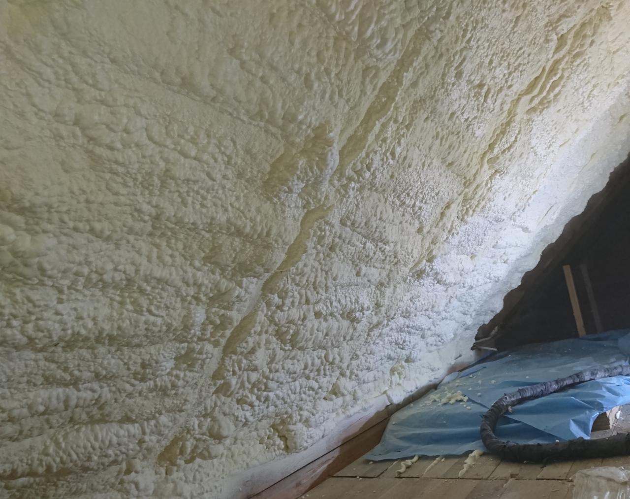 Poddasze z widoczną warstwą izolacji natryskowej, nierówna struktura piany, fragment niebieskiej folii ochronnej i wąż roboczy na podłodze.