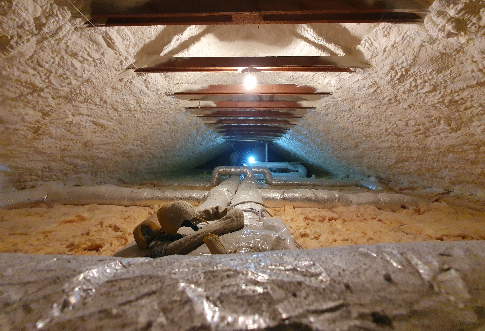 Poddasze ocieplone pianką, widoczne rury wentylacyjne, oświetlenie halogenowe i elementy konstrukcji drewnianej dachu.