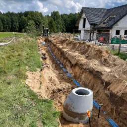 Wykop z ułożonymi niebieskimi rurami, betonowy krąg studni, łopaty i koparka w tle na budowie domu jednorodzinnego w otoczeniu zieleni.