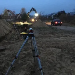Niwelator optyczny na trójnogu ustawiony na placu budowy, w tle wykop, pracująca koparka oświetlona reflektorami i samochód na drodze gruntowej. W tle zabudowa jednorodzinna o zmierzchu.