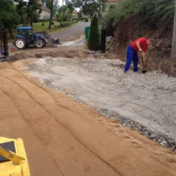 Prace przygotowawcze do budowy drogi dojazdowej: wyr&oacute;wnywanie terenu, ubijanie ziemi, rozsypywanie kruszywa, widoczny traktor z łyżką i pracownik z łopatą.