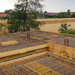 Zbrojenie stropu na budowie domu jednorodzinnego z widokiem na pola i zabudowania w tle, drewniane szalunki.