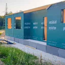 Budowa domu w stanie surowym otwartym z widoczną konstrukcją szkieletową i membraną wiatroizolacyjną z nadrukowanym logo producenta, na tle łąki i słupa wysokiego napięcia.