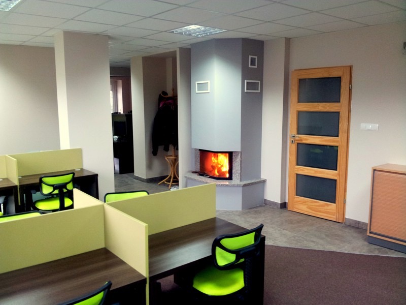 Nowoczesne biuro z zielonymi krzesłami i biurkami typu open space, z kominkiem w rogu pomieszczenia.