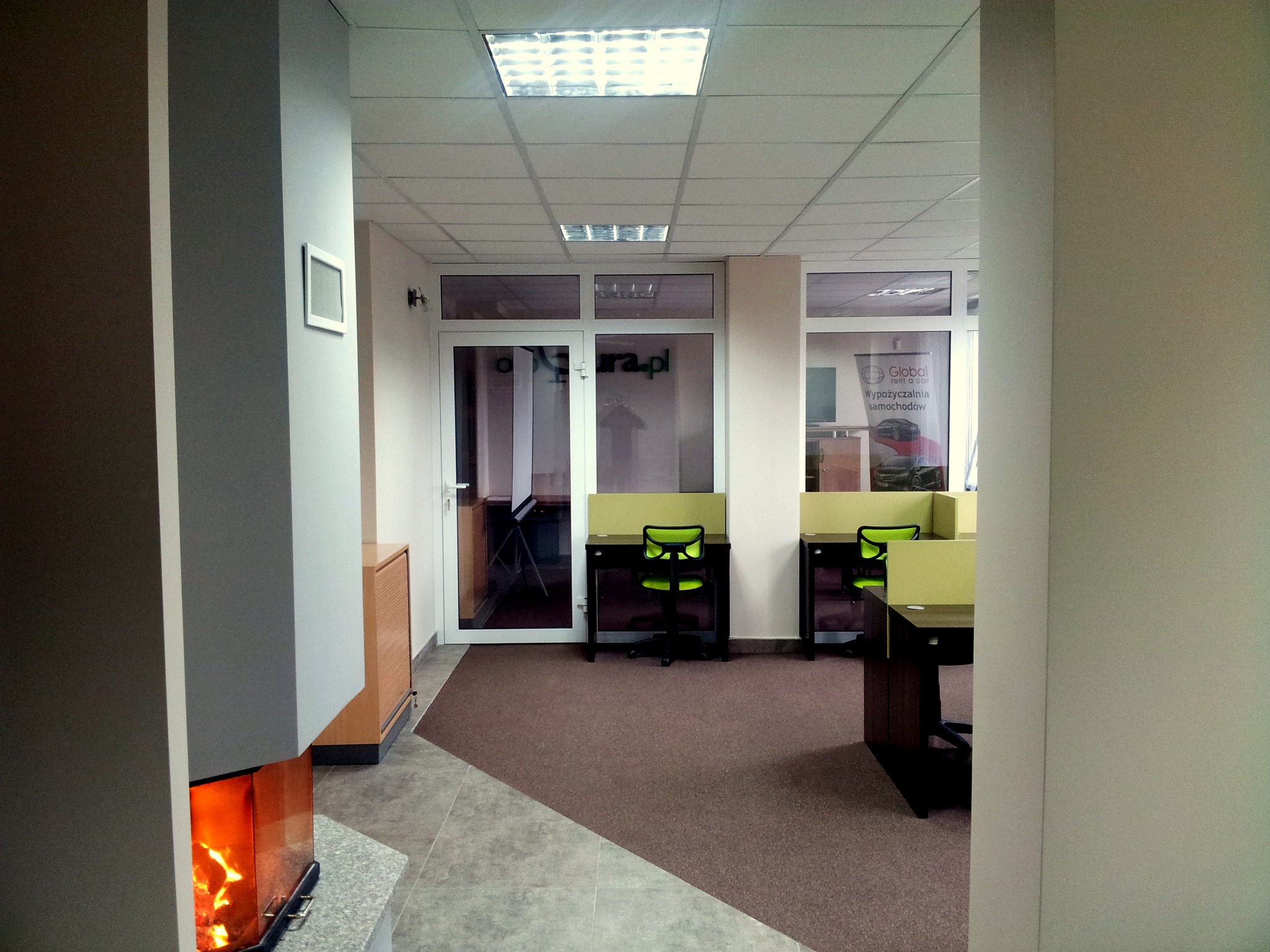 Nowoczesne wnętrze biurowe z biurkami i krzesłami w odcieniach zieleni, widoczne przez korytarz z kominkiem po lewej stronie.