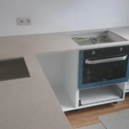 Montaż beżowego blatu kuchennego z wycięciem na zlew i płytę grzewczą, zabudowa piekarnika w trakcie instalacji, widoczne kable i elementy montażowe.