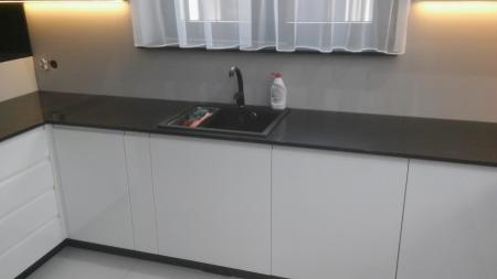 Nowoczesna kuchnia z białymi szafkami i czarnym blatem, widoczny zlew i bateria, oświetlenie LED pod szafkami wiszącymi, minimalistyczny styl.