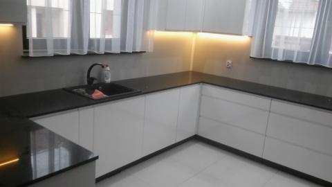 Nowoczesna kuchnia z białymi, minimalistycznymi szafkami i czarnym blatem, oświetlenie LED podszafkowe, widok na okno z białymi firankami oraz zlewozmywak z baterią.