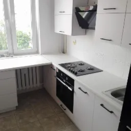 Biała kuchnia z czarnymi uchwytami, płyta gazowa i piekarnik pod oknem, widok na zieleń za oknem.