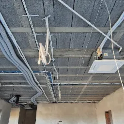 Instalacja przewodów wentylacyjnych i klimatyzacyjnych pod betonowym stropem, widoczna czarna rura z czerwonym wnętrzem, biała jednostka sufitowa i poprowadzone kable elektryczne.