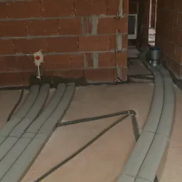 Wnętrze w trakcie budowy z widocznymi rurami wentylacyjnymi i przewodami elektrycznymi prowadzonymi po podłodze wzdłuż ścian z czerwonej cegły.