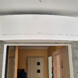 Biała jednostka wewnętrzna klimatyzatora Daikin zamontowana na szarej ścianie o fakturze betonu, widok z dołu na otwarte drzwi do pokoju z żółtymi ścianami.