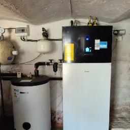 Daikin pompa ciepła z czarnym panelem sterowania i białym korpusem, obok biały zasobnik Kospel, instalacja w piwnicy z ceglaną posadzką i surowym, nierównym sufitem.