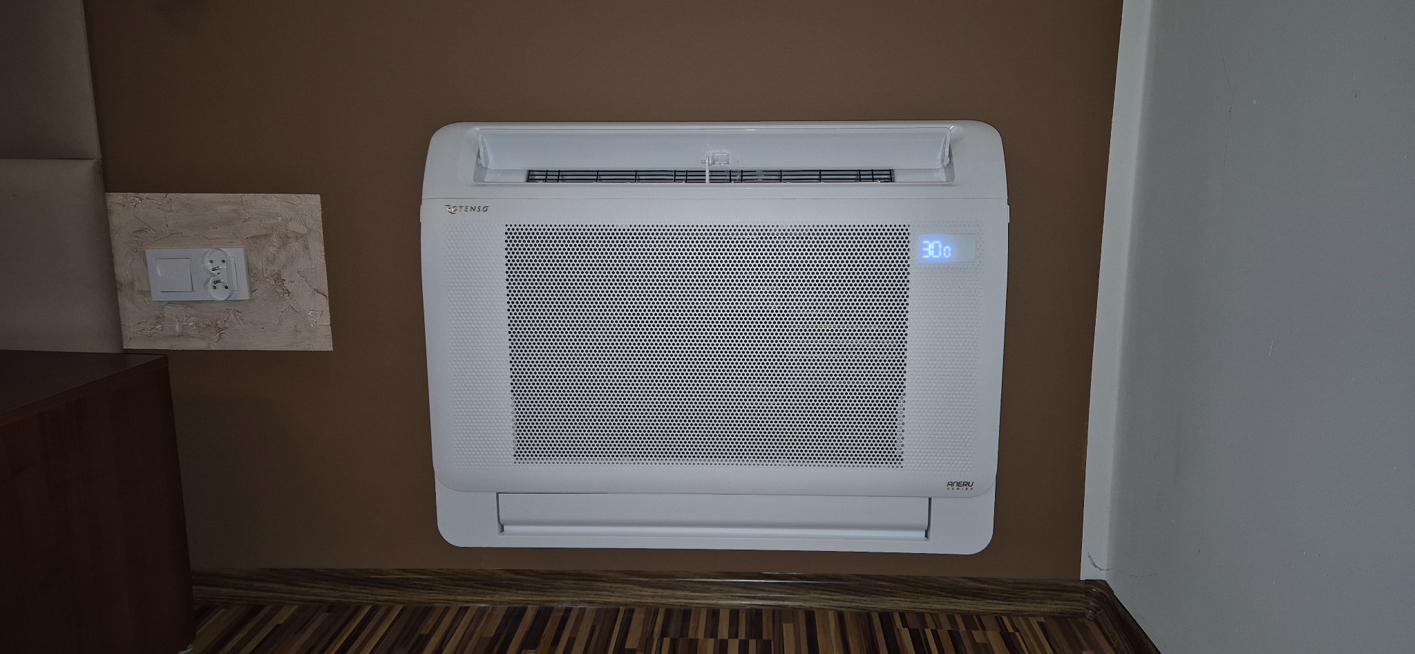 Biała jednostka klimatyzacji ściennej marki Tenso z wyświetlaczem wskazującym temperaturę 30 stopni Celsjusza, zamontowana na brązowej ścianie w pomieszczeniu z parkietem.