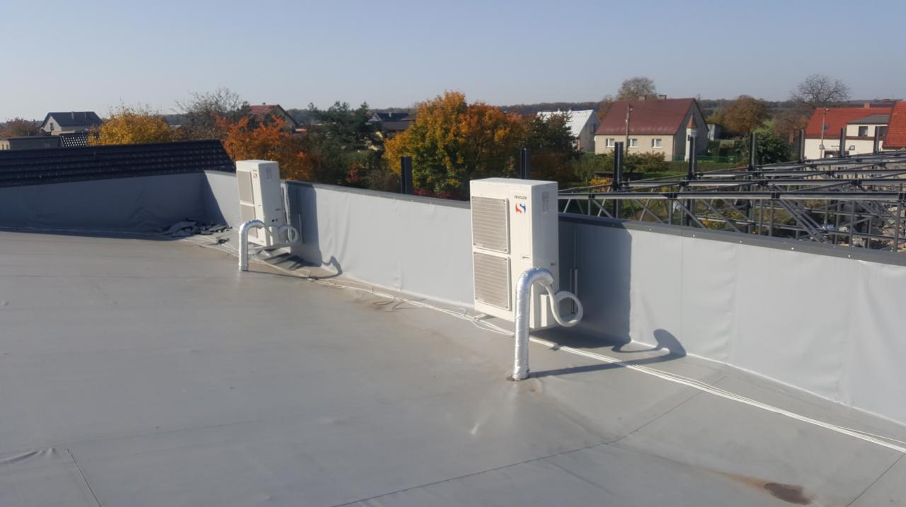 Dwa moduły zewnętrzne klimatyzacji zamontowane na płaskim dachu budynku z szarym wykończeniem, widoczne rury odprowadzające skropliny, w tle zabudowa jednorodzinna i drzewa z jesiennymi liśćmi.