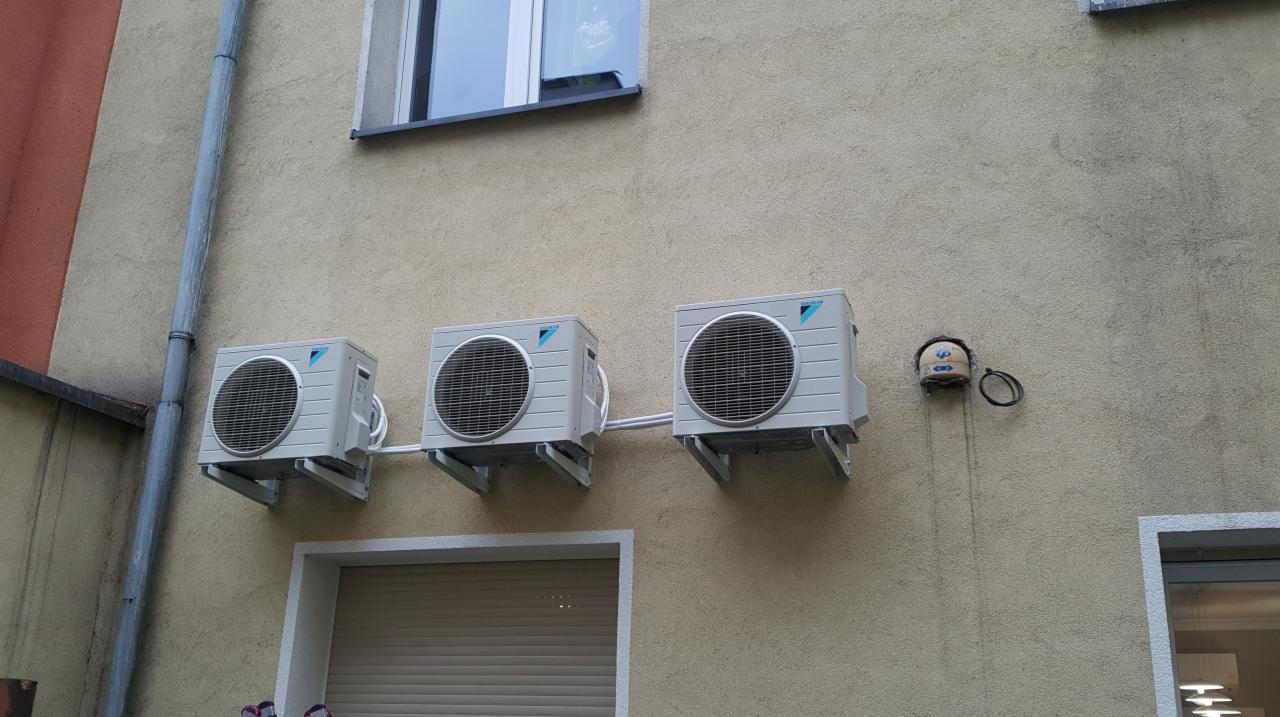 Trzy jednostki zewnętrzne klimatyzacji Daikin zamontowane szeregowo na elewacji budynku, widoczne rury odprowadzające skropliny, obok okno z refleksami.