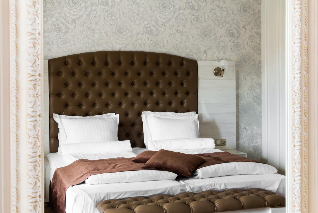 Eleganckie łóżko z brązowym, pikowanym zagłówkiem i podnóżkiem, białą pościelą i dekoracyjnymi poduszkami, widoczne przez ramę lustra w tle ściana w kwiaty.