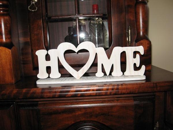 Drewniany napis HOME z sercem zamiast litery O, umieszczony na ciemnym, lakierowanym kredensie z przeszkloną nadstawką.
