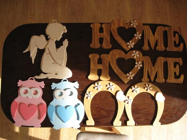 Drewniane dekoracje: aniołek, napisy HOME z sercami, sowy z sercami i podkowy z kwiatkami, leżące na ciemnobrązowym stole.