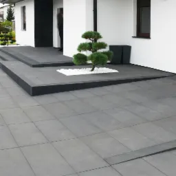 Nowoczesne szare płyty chodnikowe ułożone przed wejściem do białego domu z czarnymi akcentami, ozdobny krzew bonsai otoczony białymi kamieniami, minimalistyczne donice.