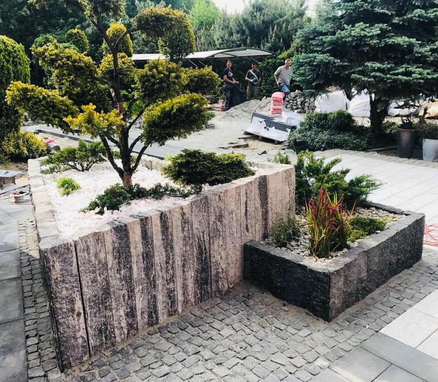 Ogród z kostki brukowej z podwyższonymi rabatami z kamienia, drzewko bonsai i białe kamyczki dekoracyjne, w tle pracownicy budowlani.