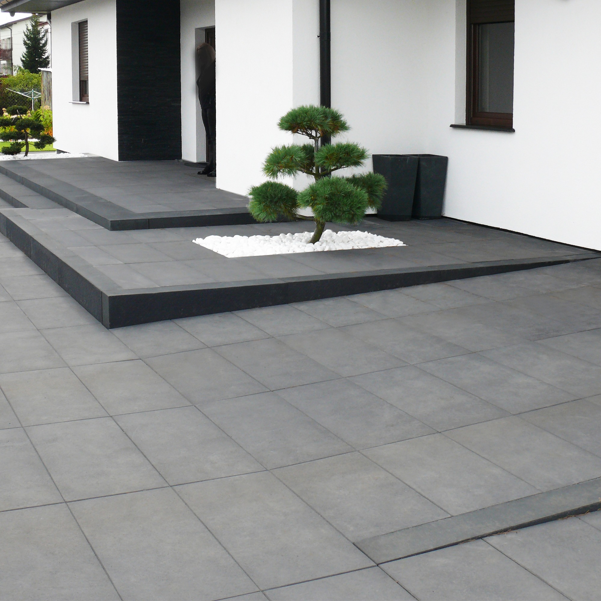 Nowoczesne szare płyty chodnikowe ułożone przed wejściem do białego domu z czarnymi akcentami, ozdobny krzew bonsai otoczony białymi kamieniami, minimalistyczne donice.