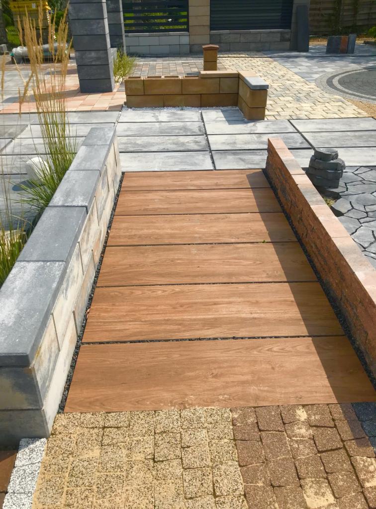 Kompozycja różnych rodzajów nawierzchni: drewnopodobne deski ułożone pomiędzy murkami z betonowych i ceglanych elementów, otoczone kostką brukową w różnych kolorach i fakturach.