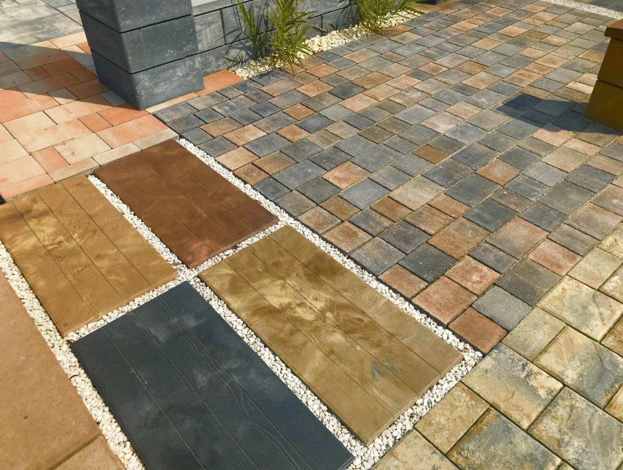 Prezentacja różnych wzorów i kolorów kostki brukowej oraz płyt imitujących drewno, oddzielonych białym grysem, na ekspozycji zewnętrznej.