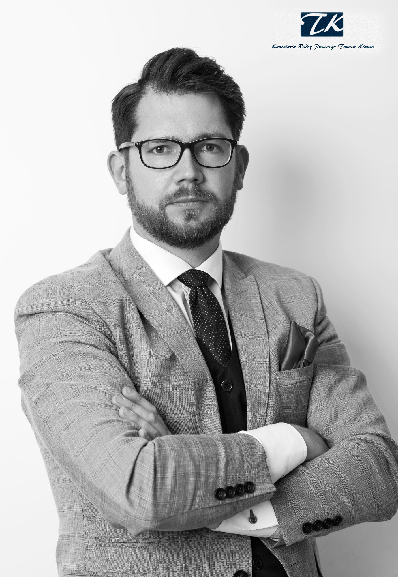 Portret w czerni i bieli: mężczyzna w okularach i garniturze w kratę, z założonymi rękami, patrzący wprost.