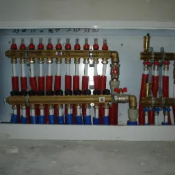 Rozdzielacz ogrzewania podłogowego w metalowej szafce, z widocznymi rurami w izolacji czerwonej i niebieskiej, oraz opisami sekcji na g&oacute;rnej listwie.