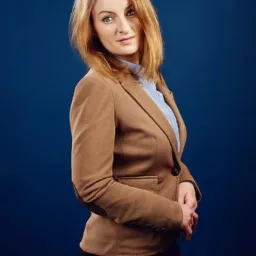 Portret kobiety w brązowej marynarce i błękitnej koszuli na ciemnoniebieskim tle, z rękami splecionymi przed sobą.