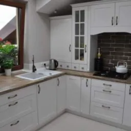 Biała kuchnia w stylu rustykalnym z drewnianym blatem, okapem nad płytą grzewczą i oknem z widokiem na zewnątrz, ozdobiona kwiatami w doniczce.