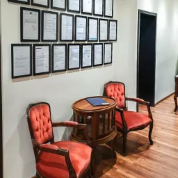 Kancelaria: Dwa czerwone, tapicerowane krzesła stoją przy drewnianym stoliku. Na ścianie wisi kilkanaście oprawionych dokumentów.