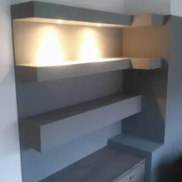 Szare, nowoczesne półki w zabudowie z oświetleniem punktowym, w tle fragment salonu z brązową kanapą.