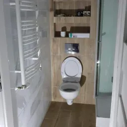 Wnętrze łazienki po remoncie z toaletą podwieszaną, grzejnikiem drabinkowym, kabiną prysznicową i płytkami imitującymi drewno na podłodze i ścianie z wnęką.