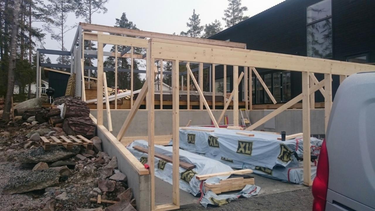 Drewniana konstrukcja rozbudowy domu w surowym stanie, widoczne materiały budowlane XL Bygg, z tyłu dom z dużymi oknami, otoczenie leśne.