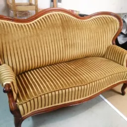 Odrestaurowana sofa w stylu retro z brązową drewnianą ramą i złocistą, prążkowaną tapicerką, widoczna w warsztacie.