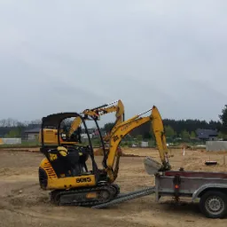 Żółta mini koparka JCB 8015 na placu budowy, obok przyczepka z narzędziami, w tle widoczne fundamenty i stosy materiałów budowlanych.