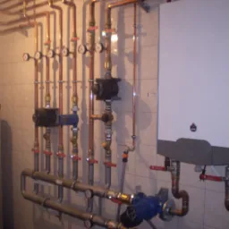 Złożona instalacja centralnego ogrzewania z kotłem gazowym, licznymi rurami miedzianymi, zaworami z czerwonymi uchwytami i manometrami, zamontowana na ścianie wyłożonej jasnymi płytkami ceramicznymi.