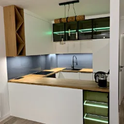 Nowoczesna kuchnia z białymi szafkami, drewnianym blatem, czarną armaturą i oświetleniem LED w zielonym kolorze, akcentowana drewnianą lampą wiszącą.