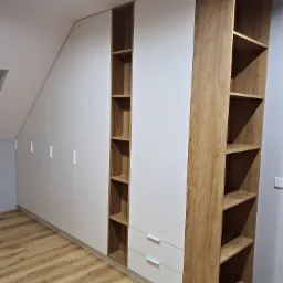 Biała szafa wnękowa z drewnianymi półkami i szufladami, dopasowana do skosu dachu, na podłodze panele imitujące drewno.