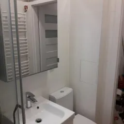 Biała łazienka z kabiną prysznicową, umywalką z szafką, toaletą i grzejnikiem drabinkowym, widoczna przez otwarte drzwi biała ściana z szarą listwą dekoracyjną.